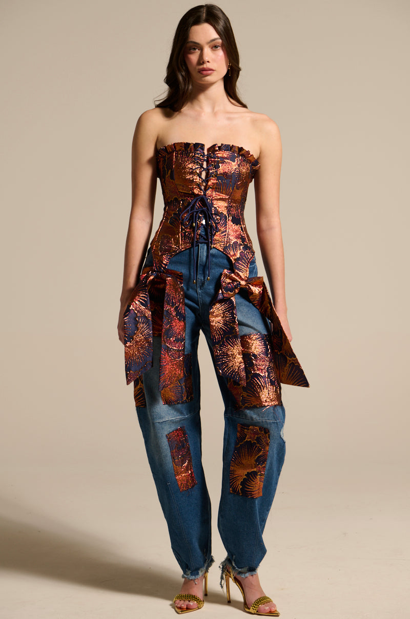 A woman with long brown hair wears ELEGANT VIBES BROCADE PATCH RELAXED DENIM, featuring brocade patches and oversized sides, paired with a strapless metallic corset and heeled sandals.