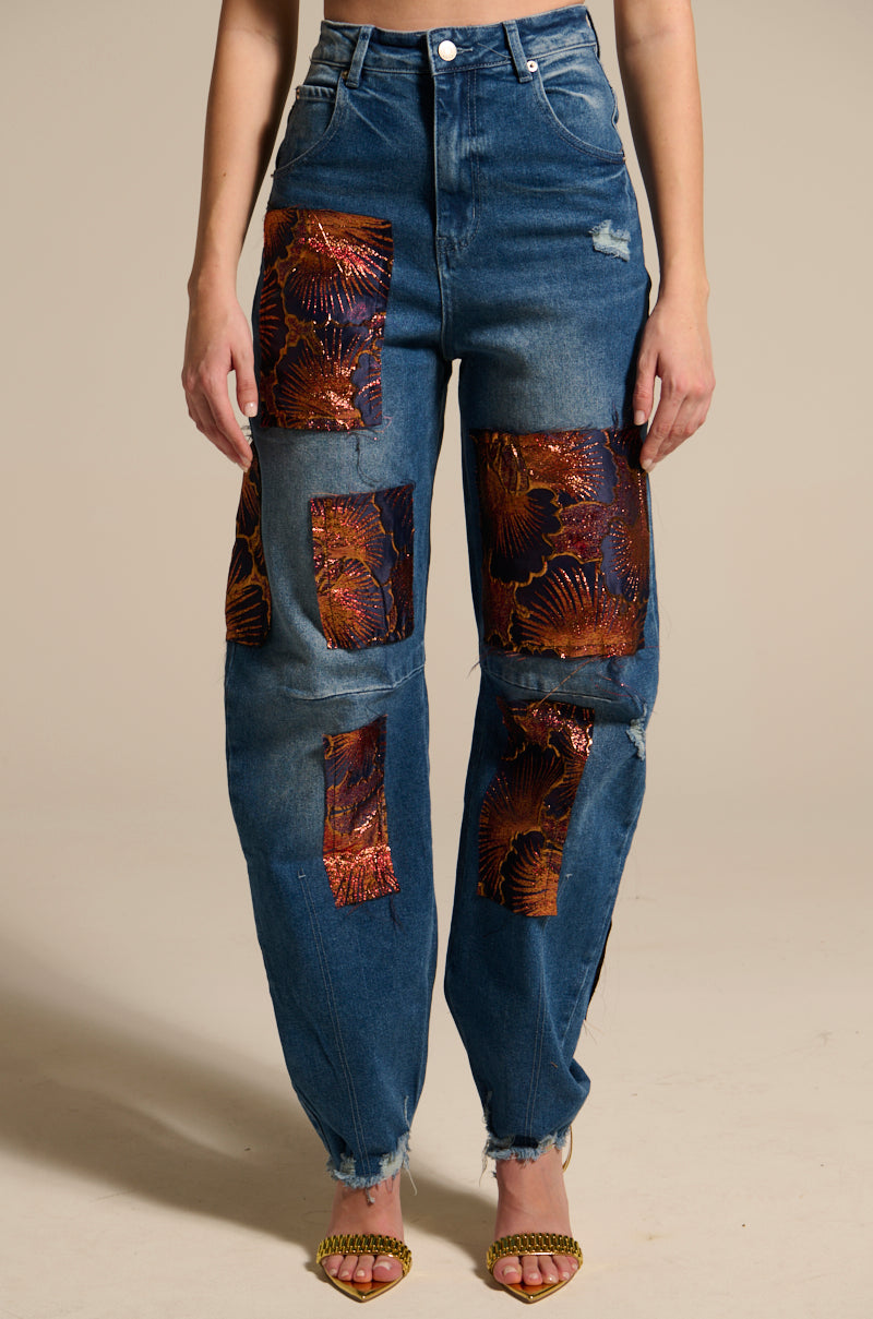 A person models the ELEGANT VIBES BROCADE PATCH RELAXED DENIM, featuring floral metallic patches and frayed hems, styled with gold open-toe heels against a plain beige background.