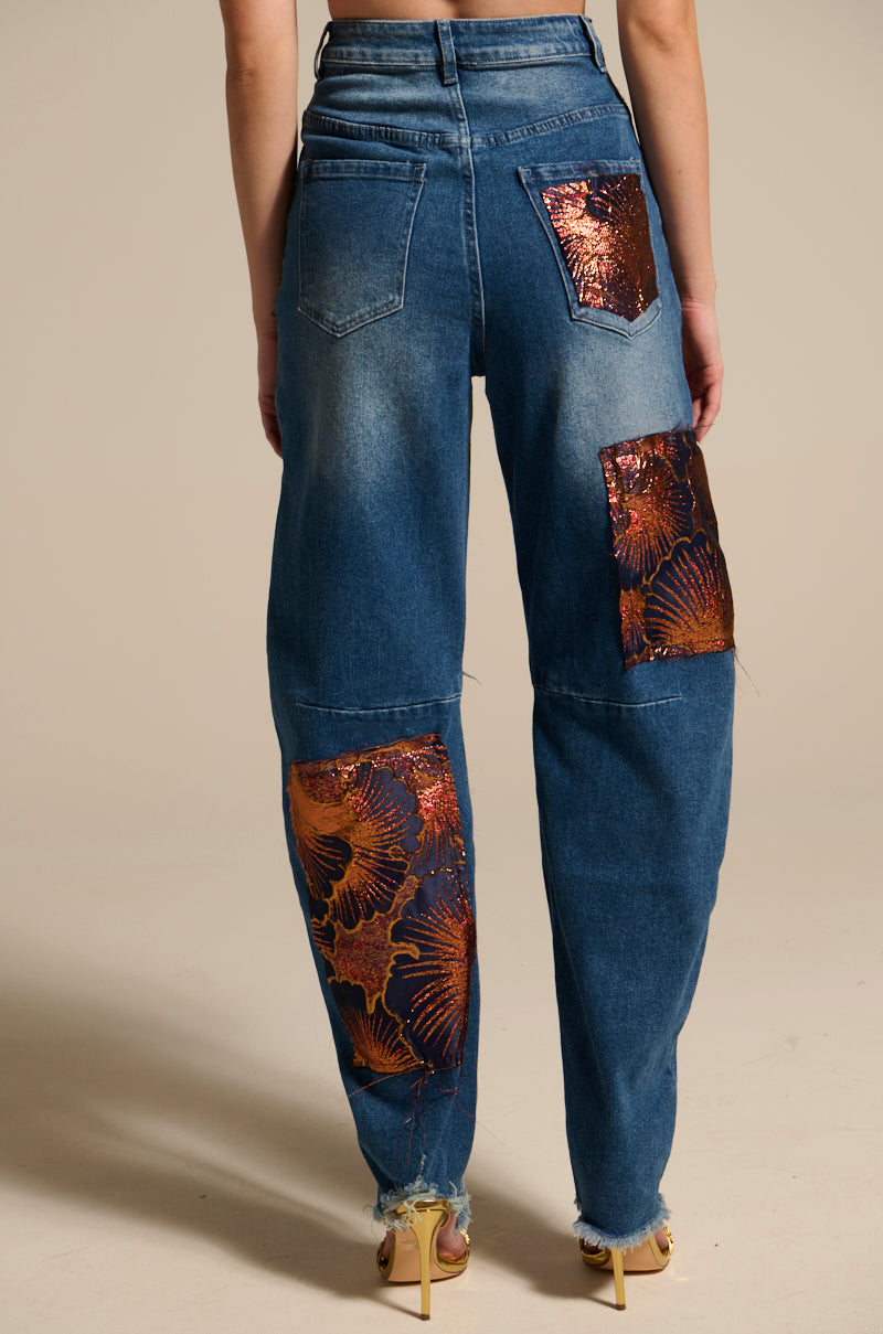Person wearing ELEGANT VIBES BROCADE PATCH RELAXED DENIM—high-rise jeans with shiny orange floral metallic patches on the back pocket, upper right leg, and lower left leg; features a faded, slightly distressed look and frayed hems.