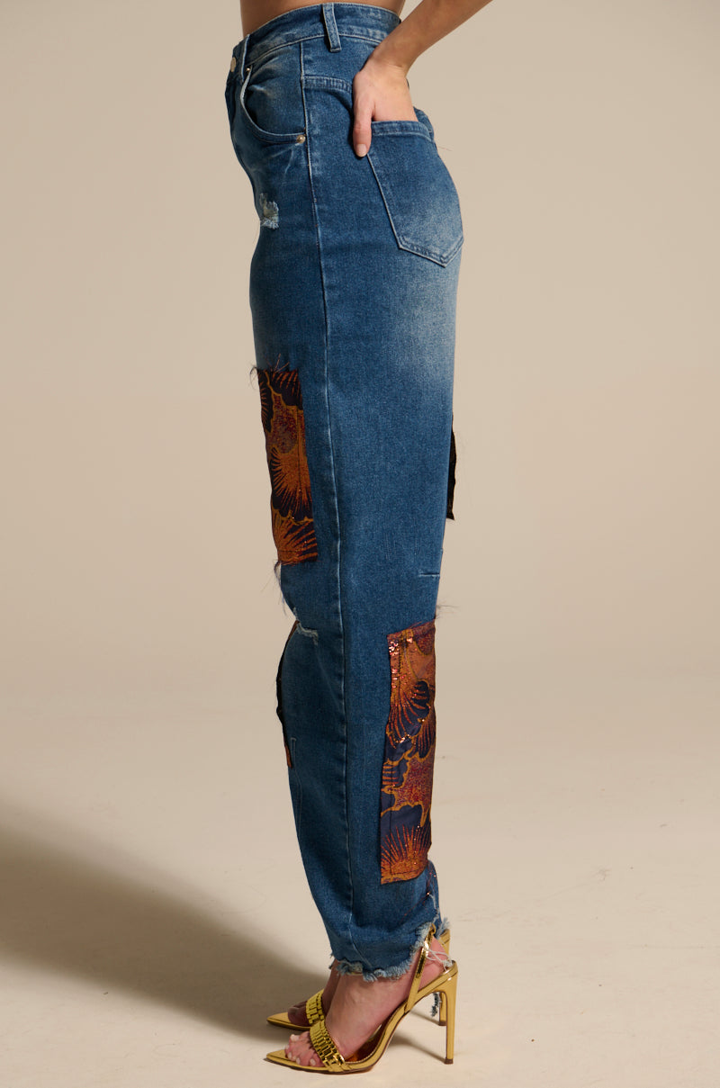 A person stands sideways in ELEGANT VIBES BROCADE PATCH RELAXED DENIM—blue high-rise jeans with brocade patches and frayed hems—paired with yellow heels, set against a plain beige background.