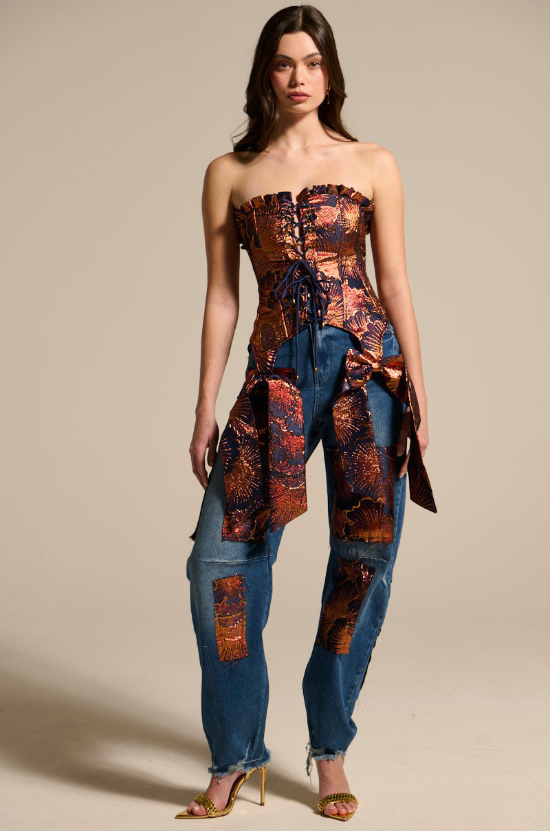 A woman models the ELEGANT VIBES BROCADE PATCH RELAXED DENIM: a strapless, metallic-patterned brocade patch corset top and high-rise jeans with floral metallic patches. She pairs it with gold heels and poses against a beige background.