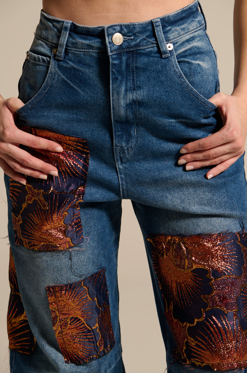 A close-up shows a person wearing ELEGANT VIBES BROCADE PATCH RELAXED DENIM, featuring high-rise design with brocade patch details and shiny red and gold floral metallic accents. Their manicured hands rest on their hips.