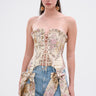 A woman wears the ROYAL ENERGY BROCADE CORSET TOP, featuring a vintage-inspired floral print, paired with high-waisted blue jeans with floral fabric panels and large bows. She has long straight hair and statement earrings against a plain white background.