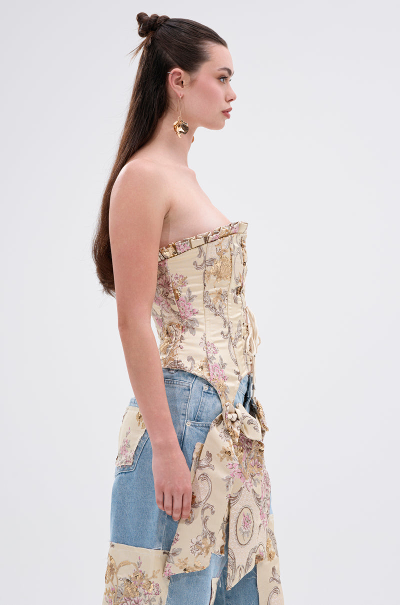 A woman with long brown hair, half tied up, stands in profile against a plain background. She wears the ROYAL ENERGY BROCADE CORSET TOP, a patchwork denim and fabric skirt, and gold earrings shaped like faces.