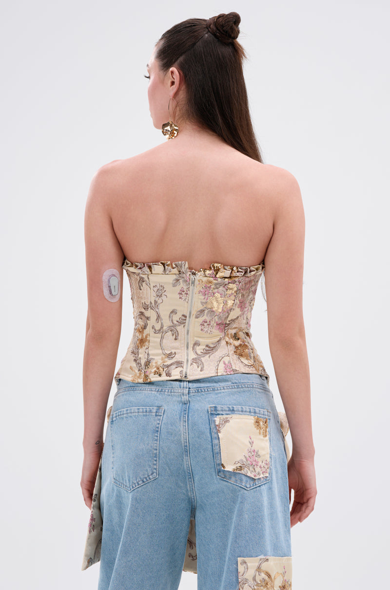 A woman with long brown hair in a half-up bun is seen from the back, wearing the ROYAL ENERGY BROCADE CORSET TOP and patchwork jeans. A medical sensor is attached to her upper left arm against a plain, light-colored background.