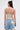 A woman with long brown hair in a half-up bun is seen from the back, wearing the ROYAL ENERGY BROCADE CORSET TOP and patchwork jeans. A medical sensor is attached to her upper left arm against a plain, light-colored background.
