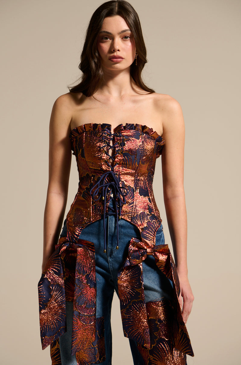 A woman models the ELEGANT VIBES BROCADE CORSET TOP—strapless with metallic floral motifs, lace-up front, ruffled trim, and matching bows—styled over denim pants against a plain beige background.