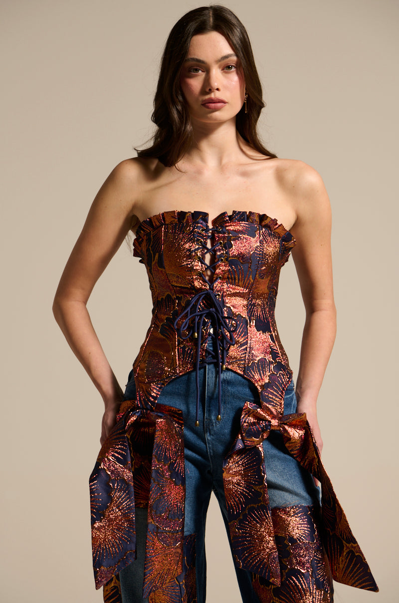 A woman with long brown hair and a neutral expression faces forward, wearing the ELEGANT VIBES BROCADE CORSET TOP—strapless with metallic floral motifs and lace-up front—paired with blue jeans featuring large matching metallic bows on the sides.