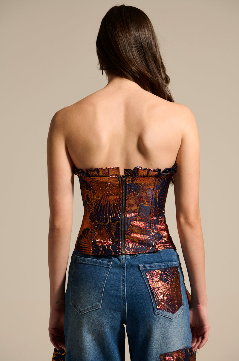 A woman with long brown hair is seen from behind wearing the ELEGANT VIBES BROCADE CORSET TOP, featuring metallic floral motifs, paired with blue jeans with metallic patches against a neutral background.