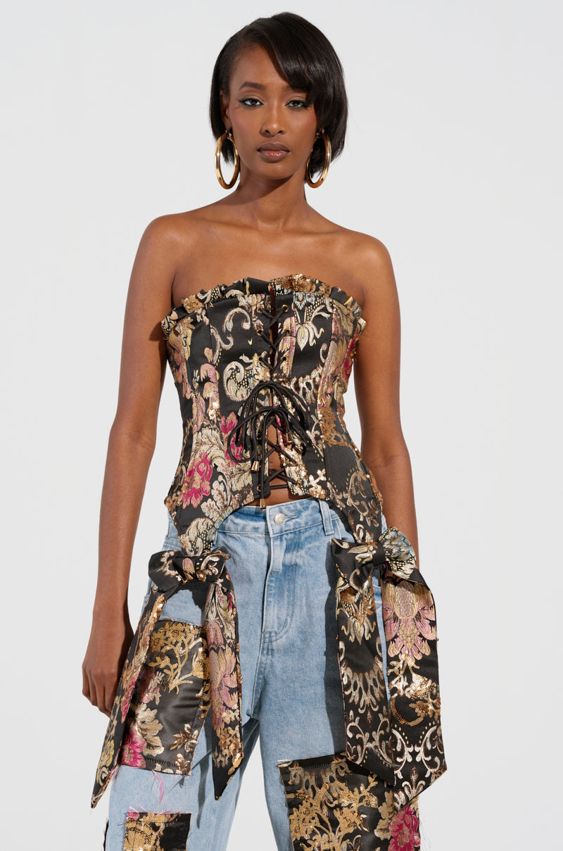 A woman exudes confidence in her QUEENDOM BROCADE CORSET TOP, featuring a stunning floral brocade pattern and a lace-up front. Her ensemble is complemented by long earrings and jeans decorated with matching floral fabric patches, all set against a plain white backdrop.