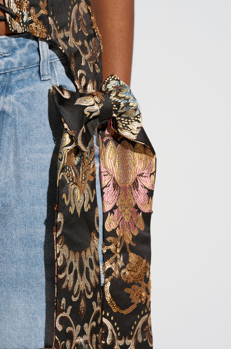 A person in light blue jeans dons a QUEENDOM BROCADE CORSET TOP with a tied bow at the waist. This garment features a black, gold, and pink floral brocade pattern, showcasing intricate designs and shimmering metallic accents.
