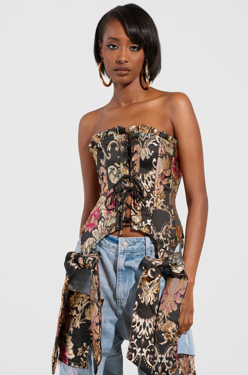 A person stands confidently in a fashionable QUEENDOM BROCADE CORSET TOP showcasing a black and gold floral brocade pattern, paired with light blue jeans. The ensemble is accentuated by prominent ribbon details and a lace-up front panel, set against a plain and minimalistic background to emphasize the outfit.