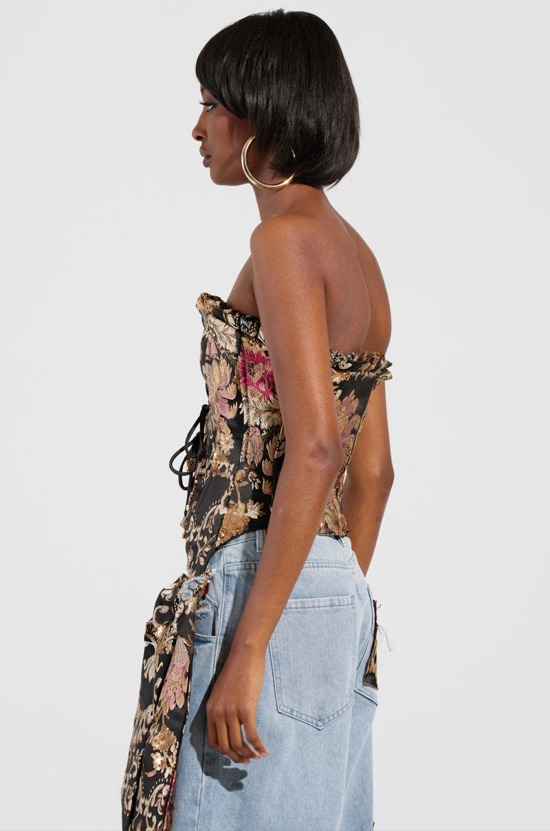 A woman with short dark hair poses in a QUEENDOM BROCADE CORSET TOP that showcases a floral brocade design and a lace-up front panel, combined with light blue denim pants. She's accessorized with large gold hoop earrings and is turned sideways against a plain background.