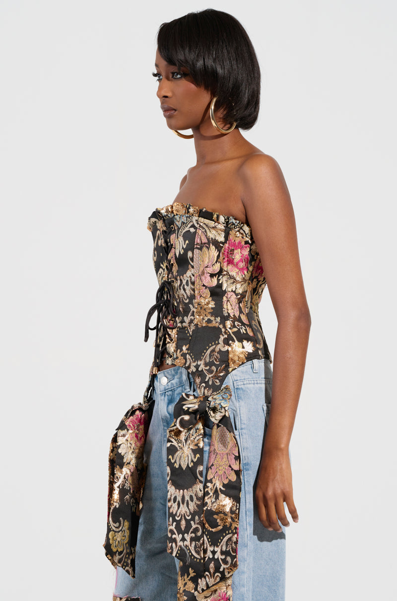 A person with short hair stands sideways, wearing a strapless QUEENDOM BROCADE CORSET TOP paired with light blue jeans. This corset showcases a lace-up front panel and features a black base enhanced with pink, gold, and green floral brocade patterns. Hoop earrings perfectly complement the chic ensemble.