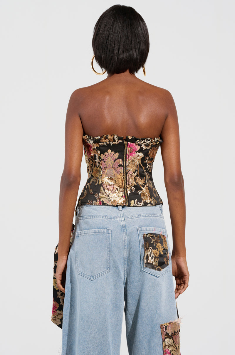 A person with short hair stands facing away, wearing the QUEENDOM BROCADE CORSET TOP paired with light blue jeans featuring floral patches. The background is plain white.