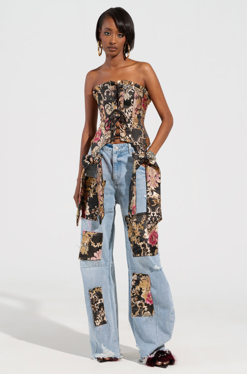 A person sporting a QUEENDOM BROCADE CORSET TOP showcasing a lace-up front panel and light blue patchwork jeans stands against a plain background. The jeans are adorned with floral patches that complement the top's floral brocade design. They exude confidence, with one hand resting on their hip.