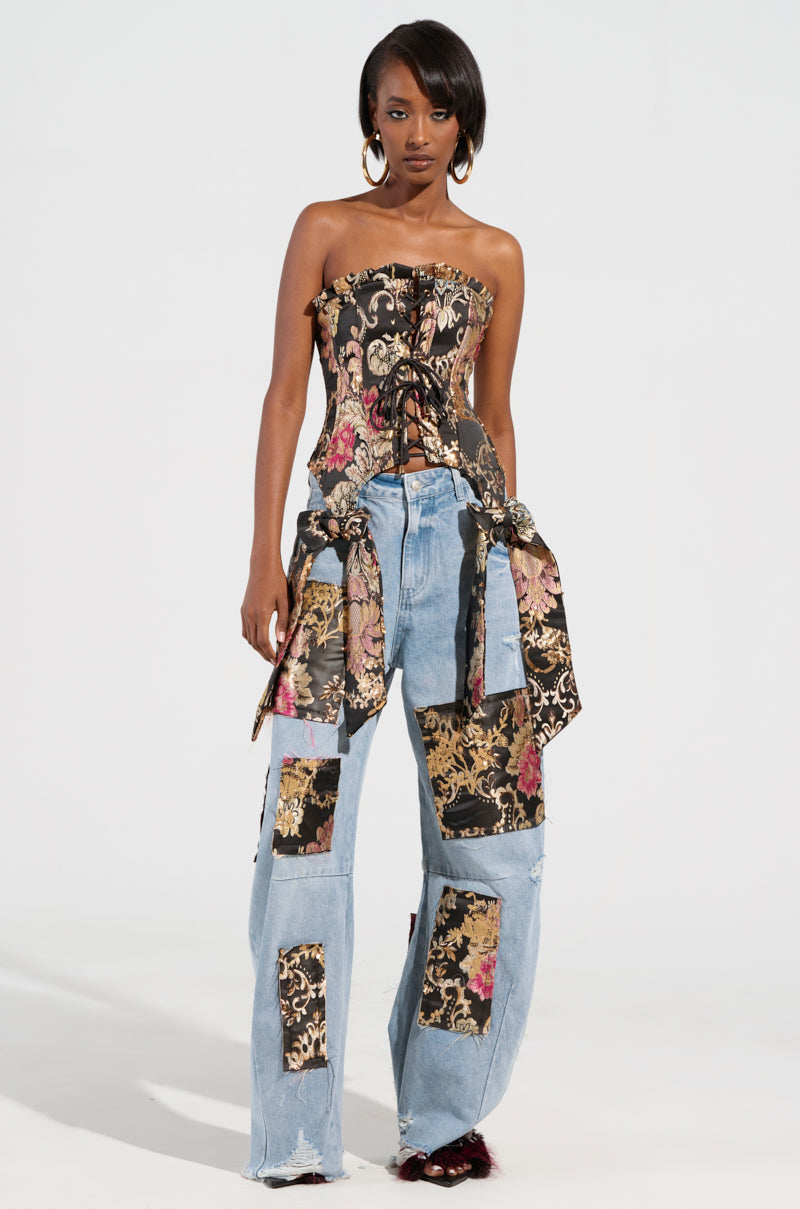 A woman wears a QUEENDOM BROCADE CORSET TOP featuring a floral brocade design, paired with blue denim jeans. The jeans are adorned with matching floral patches and have a tied fabric around the waist. Hoop earrings sparkle with metallic accents as her long, dark hair flows against a simple white backdrop.