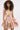A woman with curly hair wears the PRETTY IN PINK BROCADE MINI DRESS, a strapless metallic dress with a pink, gold, and green floral brocade pattern and a flared ruffled skirt, posed against a plain light background.