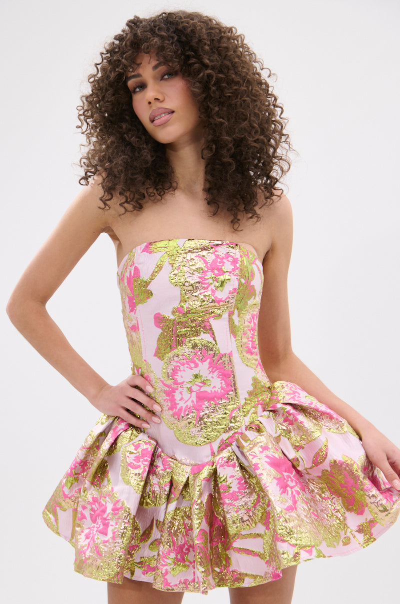 A woman with curly hair poses against a plain background wearing the PRETTY IN PINK BROCADE MINI DRESS—strapless, with a pink, gold, and white floral pattern and a flared ruffled mini skirt.