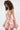 A woman with curly hair wears the PRETTY IN PINK BROCADE MINI DRESS, featuring a strapless neckline and pink-gold floral pattern. The shimmery brocade stands out as she poses against a plain light background, turned slightly to the side.