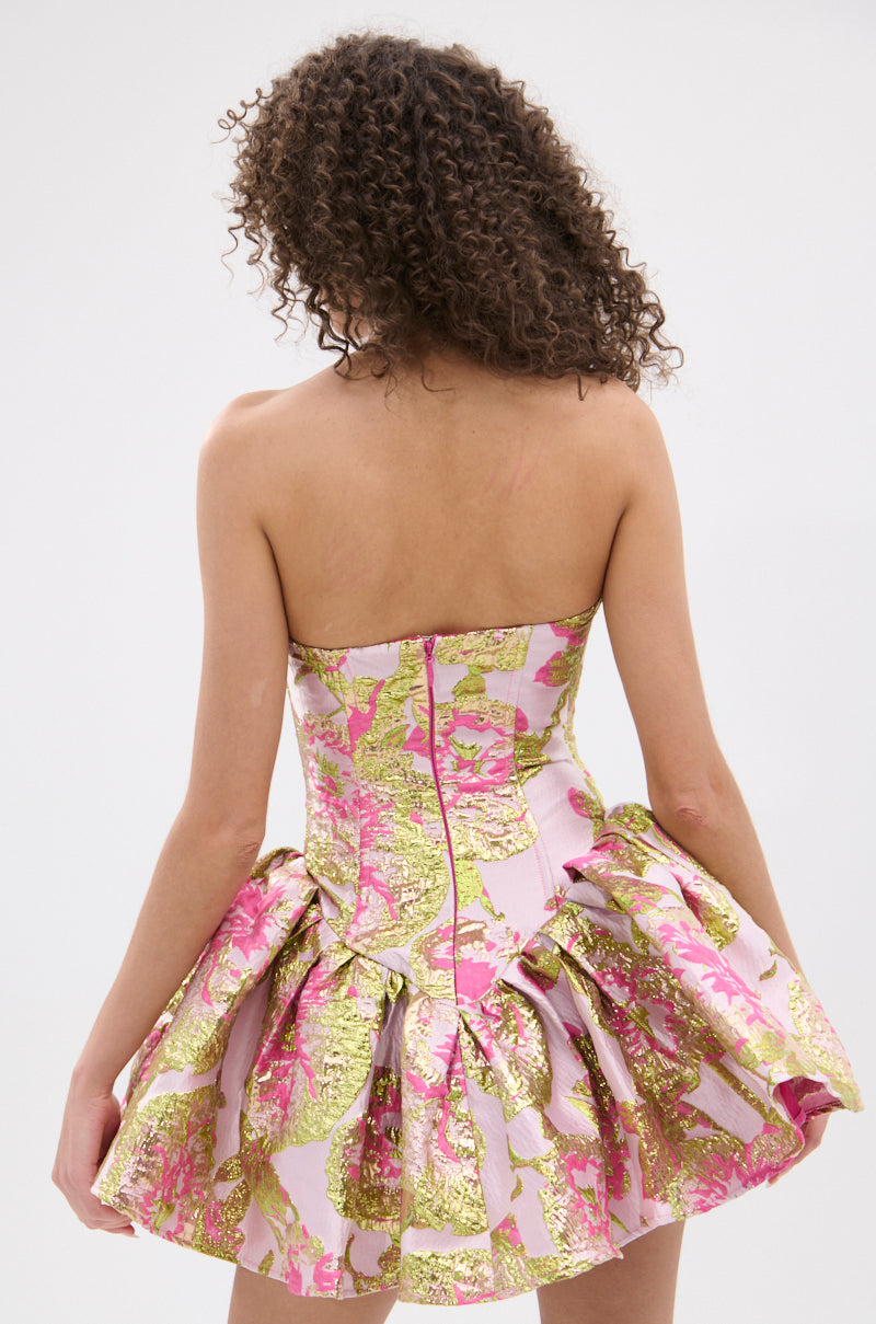 A woman with curly hair models the PRETTY IN PINK BROCADE MINI DRESS, featuring a strapless neckline and a flared, ruffled skirt. She faces away from the camera, showcasing the dress’s back against a plain white background.