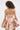 A woman with curly hair models the PRETTY IN PINK BROCADE MINI DRESS, featuring a strapless neckline and a flared, ruffled skirt. She faces away from the camera, showcasing the dress’s back against a plain white background.