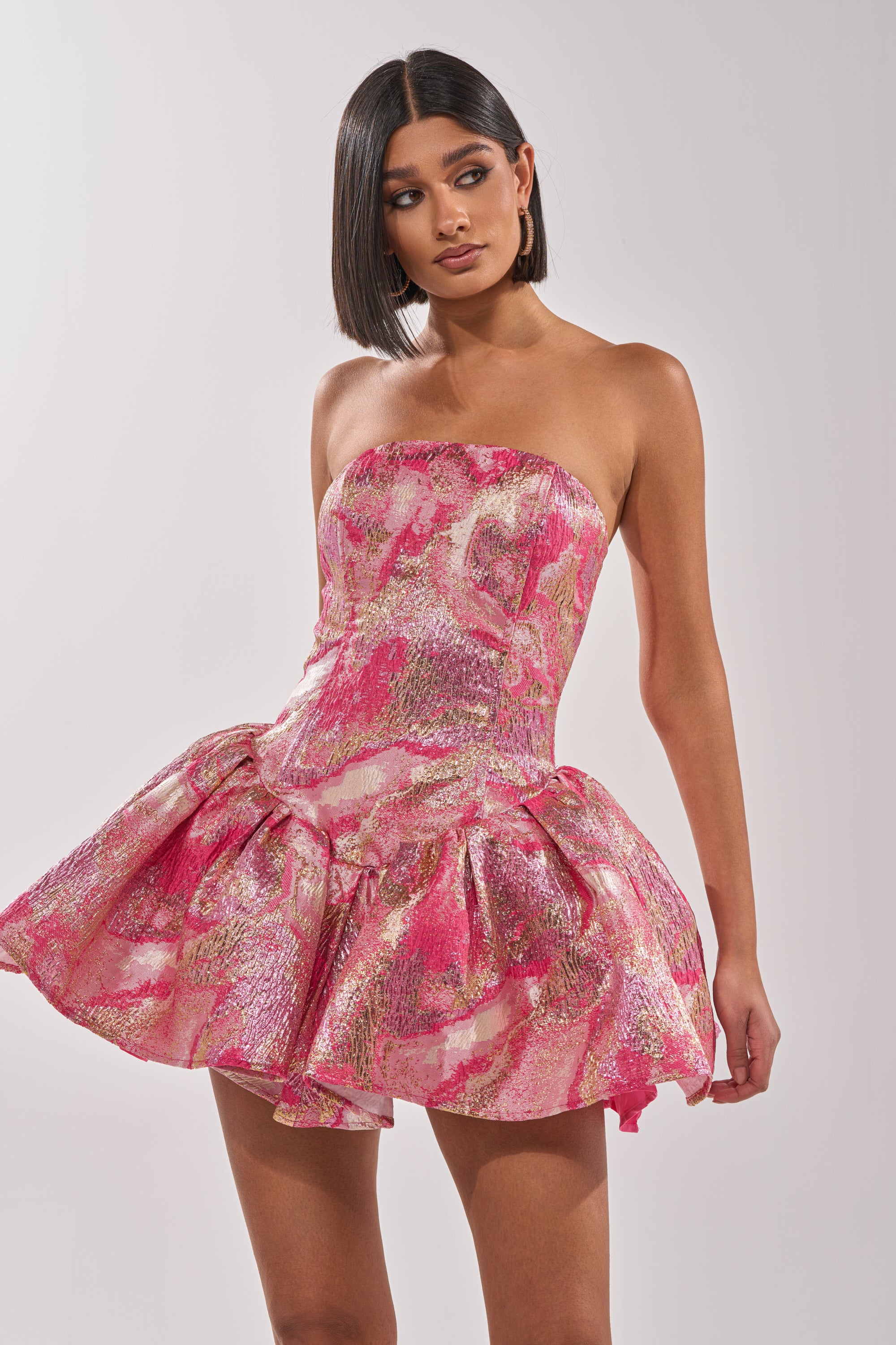 A woman wears the PRETTY IN PINK BROCADE MINI DRESS IN PINK MULTI, featuring a strapless design with pink, gold, and white abstract patterns. She stands against a plain background, looking to her left with arms relaxed at her sides. 3