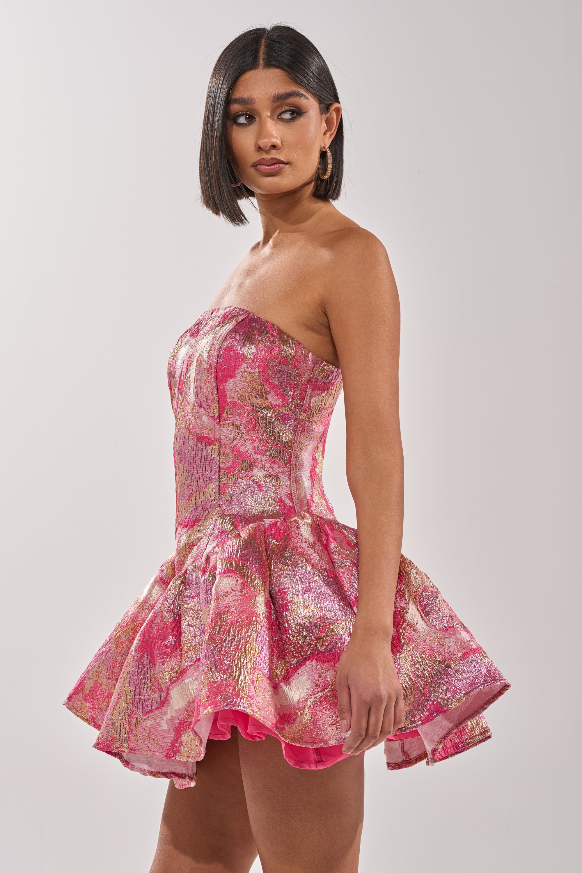 A woman with straight, shoulder-length dark hair models the PRETTY IN PINK BROCADE MINI DRESS IN PINK MULTI, featuring a strapless corset-style bodice and flared skirt, as she stands sideways against a plain background. 4