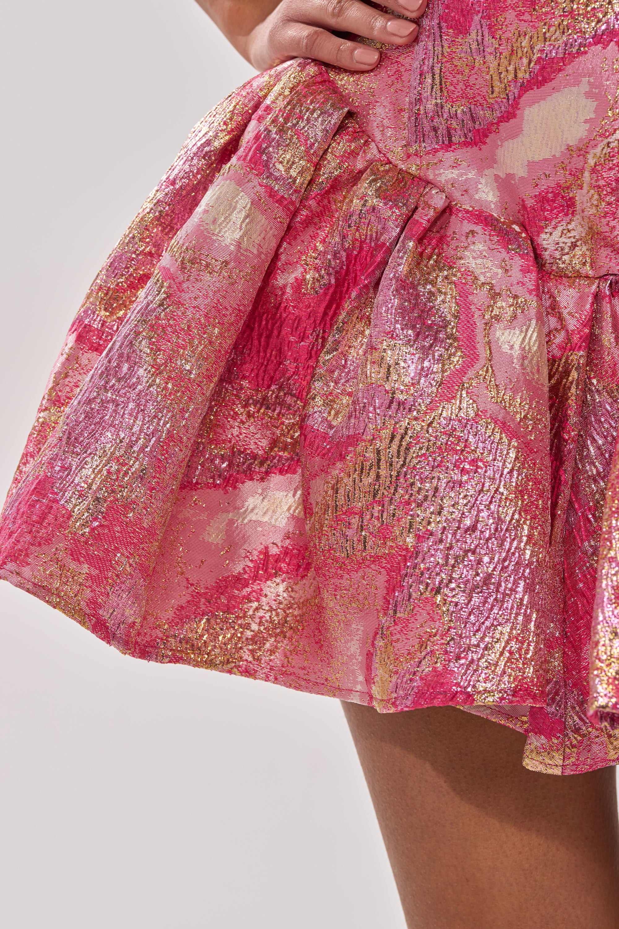 A close-up of a woman's hand resting on the waist of the PRETTY IN PINK BROCADE MINI DRESS IN PINK MULTI, featuring a strapless corset design, flared skirt, shimmering abstract patterns, and textured brocade for a glamorous look. 7