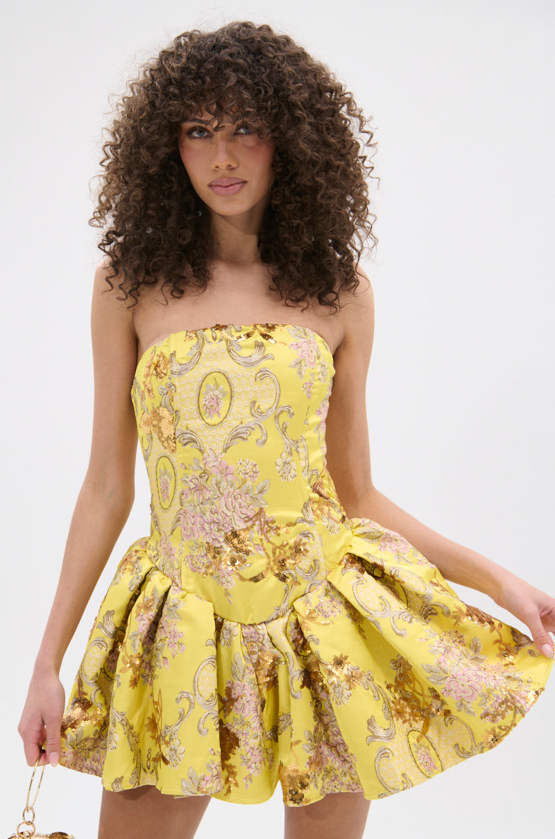 A woman with curly hair models the CRAZY IN LOVE BROCADE MINI DRESS, a strapless mini with metallic embroidery and pink and gold floral patterns. She stands against a plain white background, holding the flared skirt with both hands.