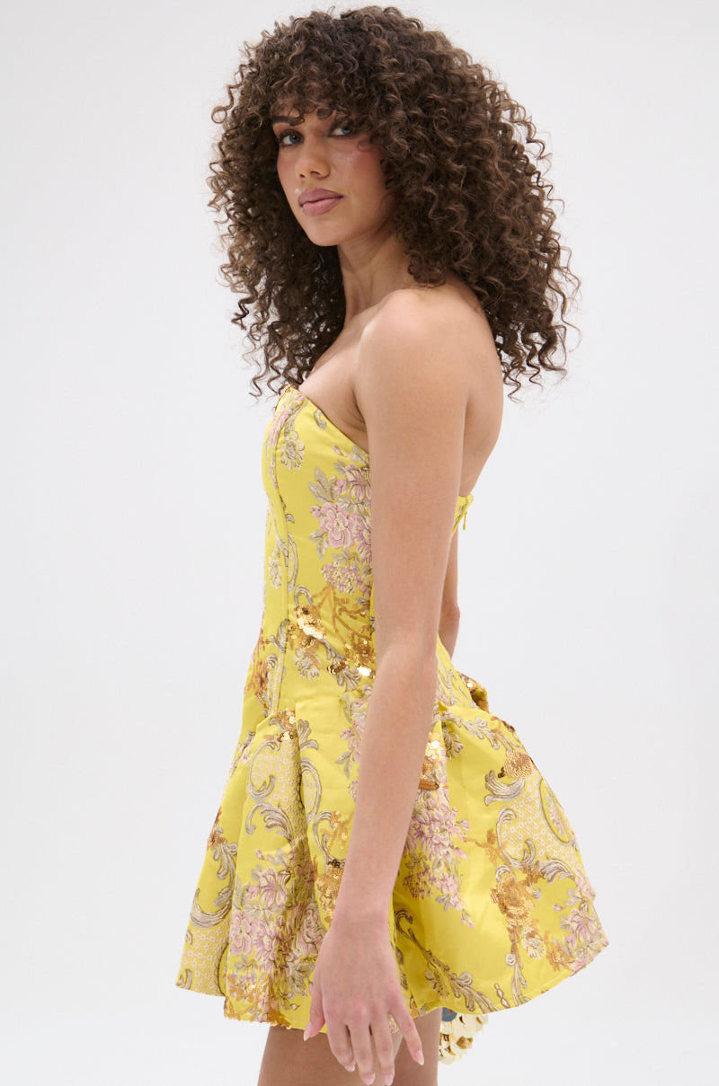 A woman with curly hair stands in profile against a white background, modeling the yellow, strapless "CRAZY IN LOVE BROCADE MINI DRESS.