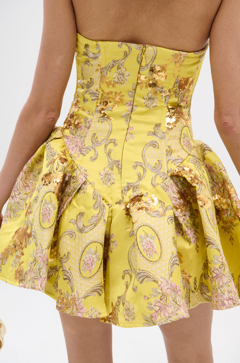Close-up of a person wearing the CRAZY IN LOVE BROCADE MINI DRESS, featuring a strapless neckline, yellow brocade fabric with gold and pink floral patterns, metallic embroidery, sequins, structured bodice, and flared skirt.