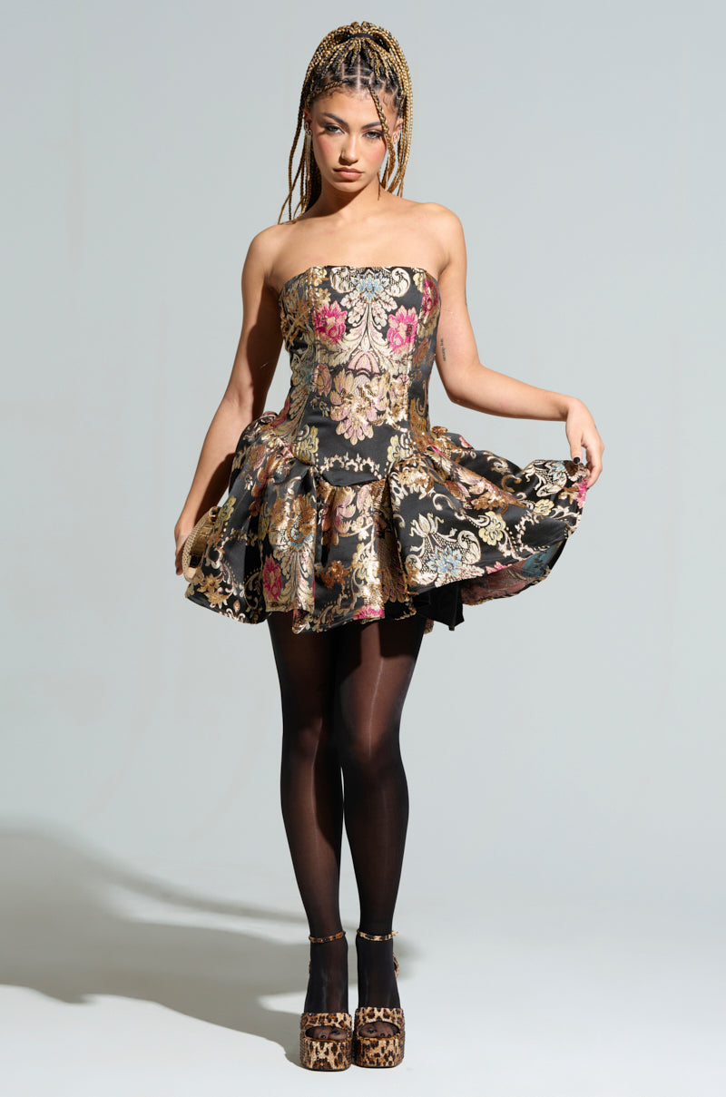 A person with braided hair poses confidently, showcasing their PURE ROYALTY BROCADE MINI DRESS. They hold the sides of its strapless, floral-patterned design, wearing black tights and leopard-print platform shoes, standing gracefully against a plain background.