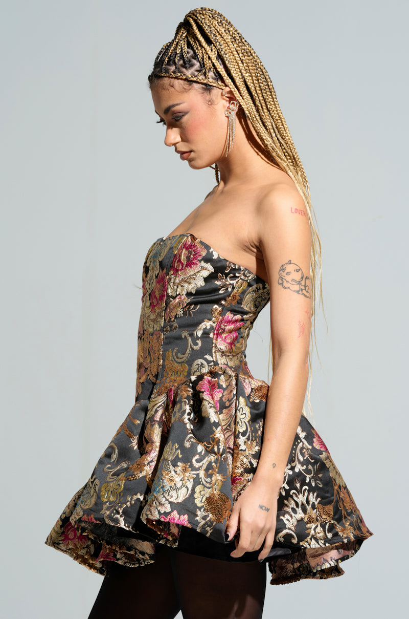 A woman with long braided hair is elegantly poised in profile against a plain background, wearing the PURE ROYALTY BROCADE MINI DRESS, which showcases delicate floral patterns and subtle metallic brocade accents. Her arms are adorned with several tattoos, and she wears delicate earrings as she gazes downward.