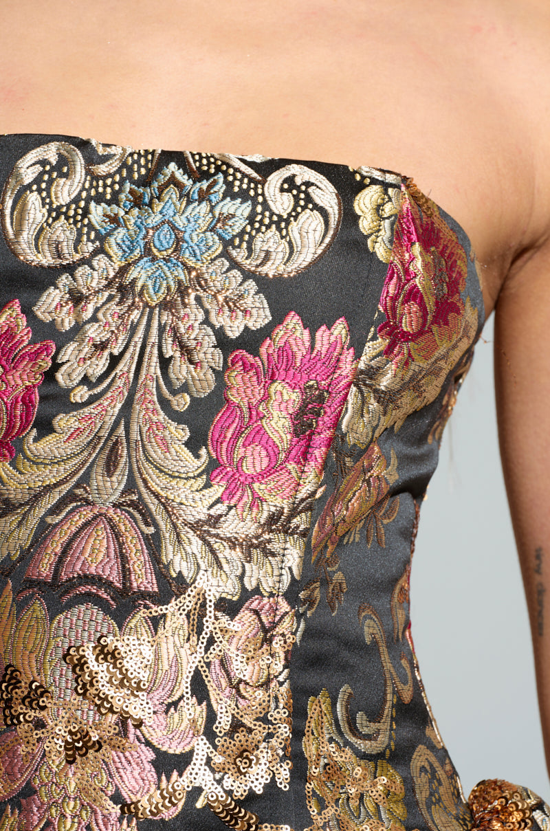 Close-up of the PURE ROYALTY BROCADE MINI DRESS, an elegant strapless design featuring a bubble skirt with an intricate floral and paisley pattern in pink, gold, and blue on a dark backdrop. A subtle hint of a tattoo can be seen on the person's arm in the background.