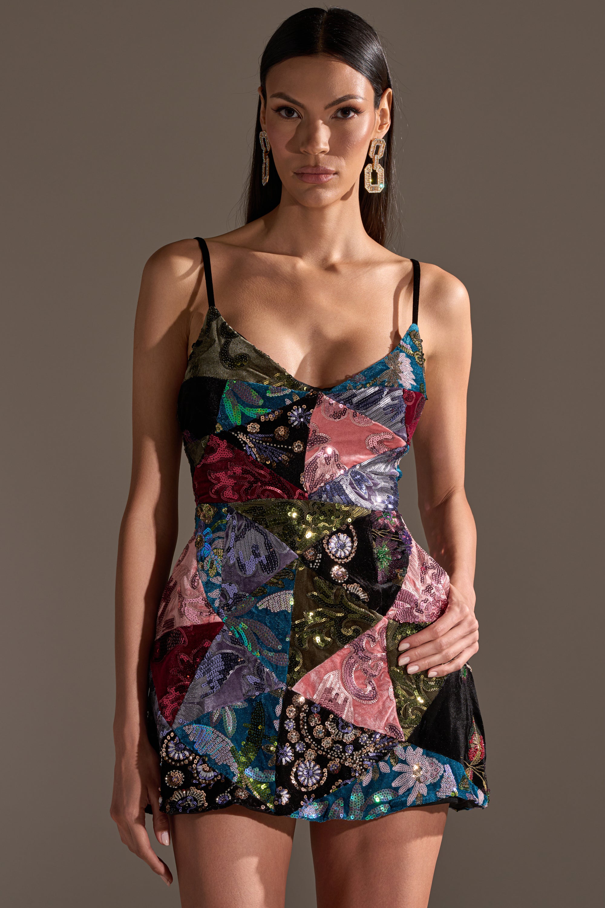 A woman stands against a plain background, wearing the MIXED EMOTIONS BROCADE MINI DRESS featuring thin straps and decorative details. With straight dark hair and bold earrings, she showcases the vibrant mini dress’s unique patchwork style.