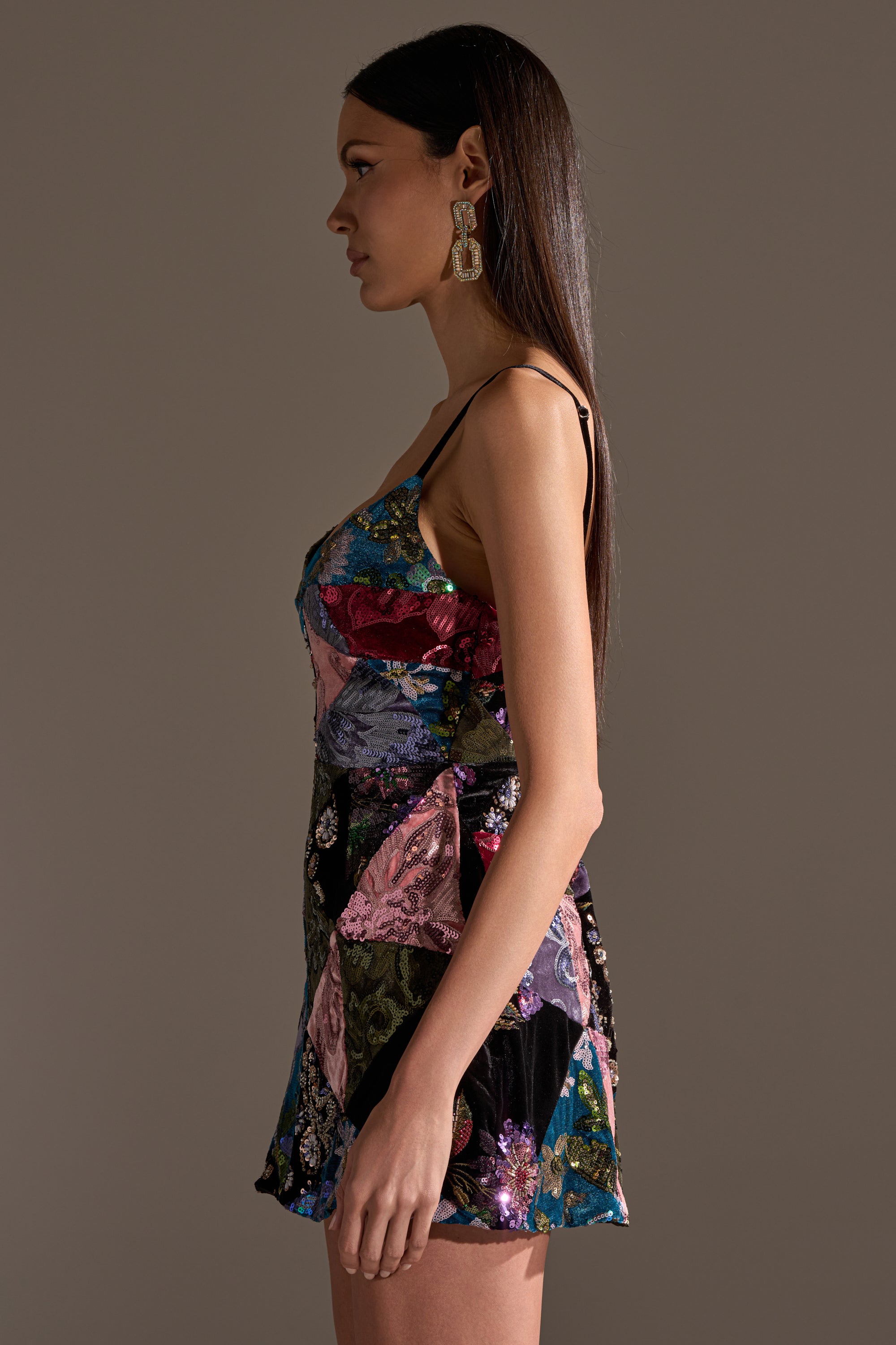A woman in profile wears the MIXED EMOTIONS BROCADE MINI DRESS, a sleeveless fitted mini in jewel tone patchwork. She has long, straight hair and large geometric earrings, standing against a plain background.