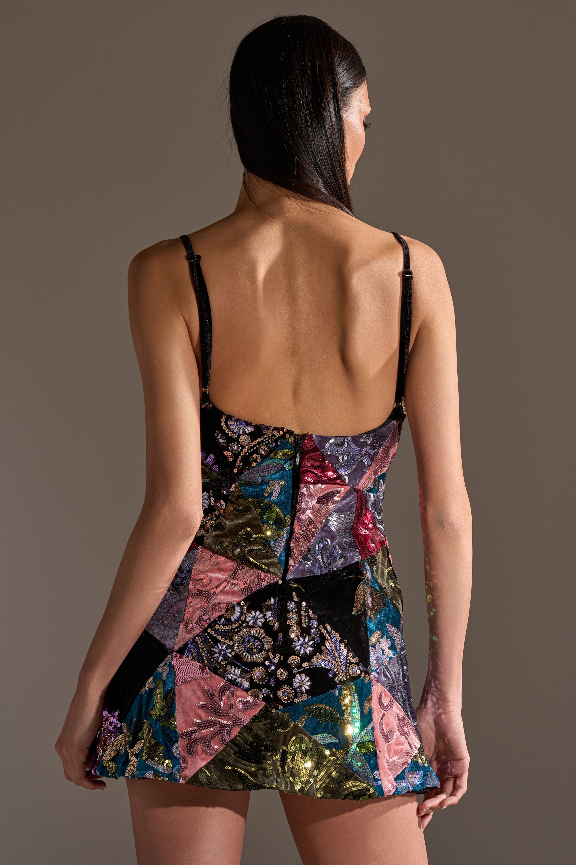 A person with long dark hair models the MIXED EMOTIONS BROCADE MINI DRESS, a sleeveless, backless mini dress with sequin floral patchwork, standing against a plain background.
