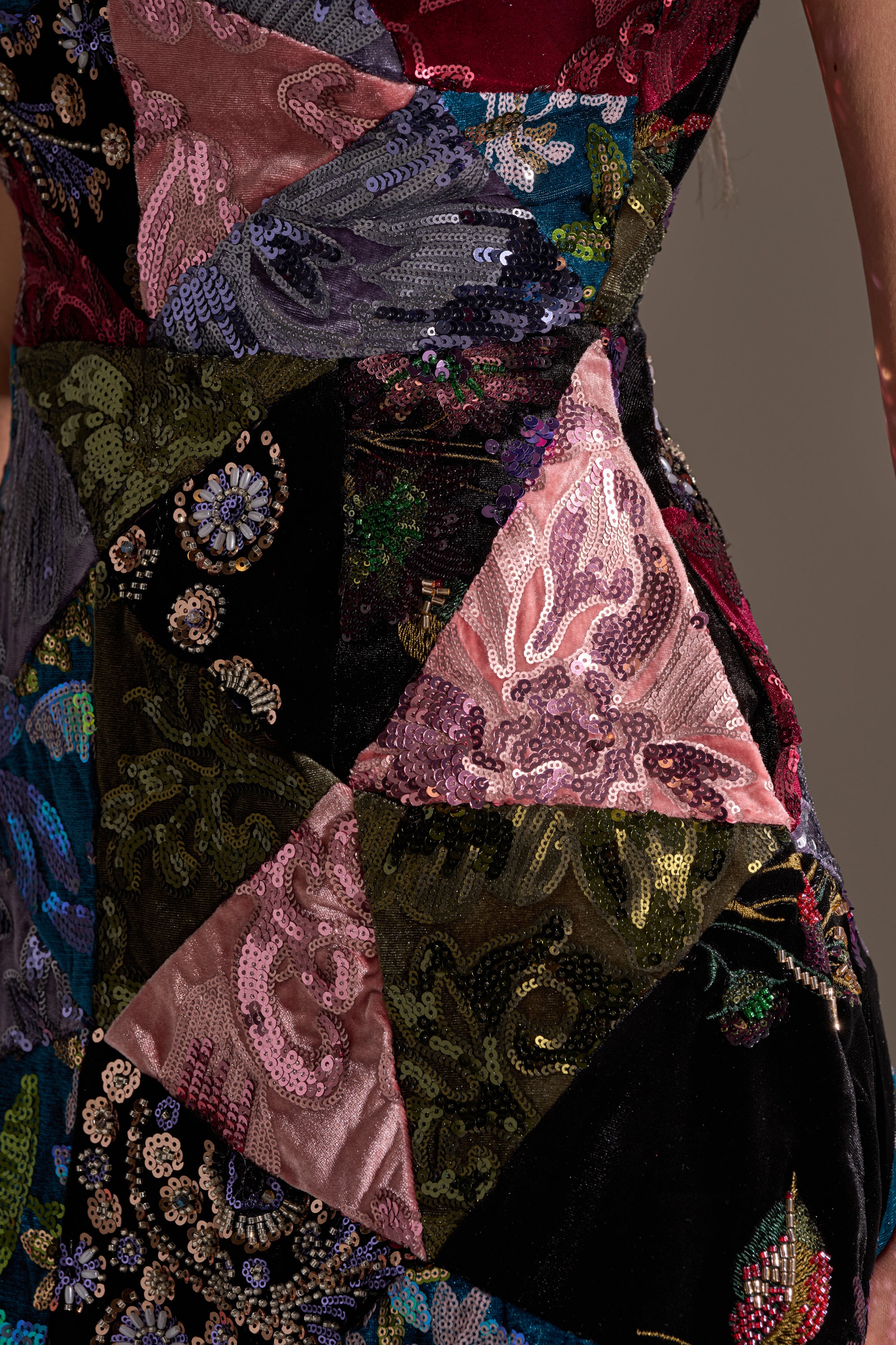 Close-up of the MIXED EMOTIONS BROCADE MINI DRESS, featuring triangular patchwork in pink, black, purple, and green with sequins and detailed embroidery. A person's arm is partly shown on the right.