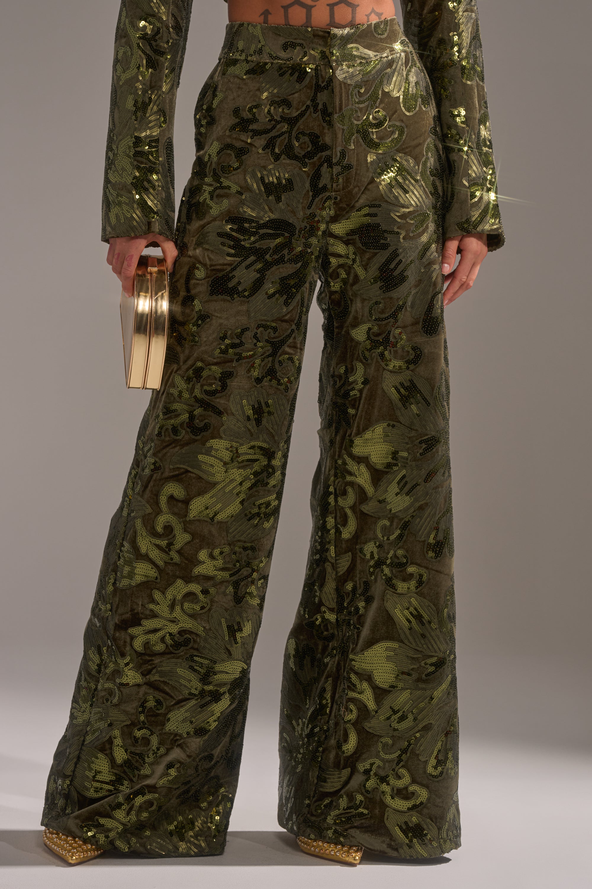 Wearing green AURORA SEQUIN VELVET TROUSER with a shiny baroque floral pattern, gold pointed-toe shoes, and holding a small gold clutch. Only the lower torso and legs are shown.