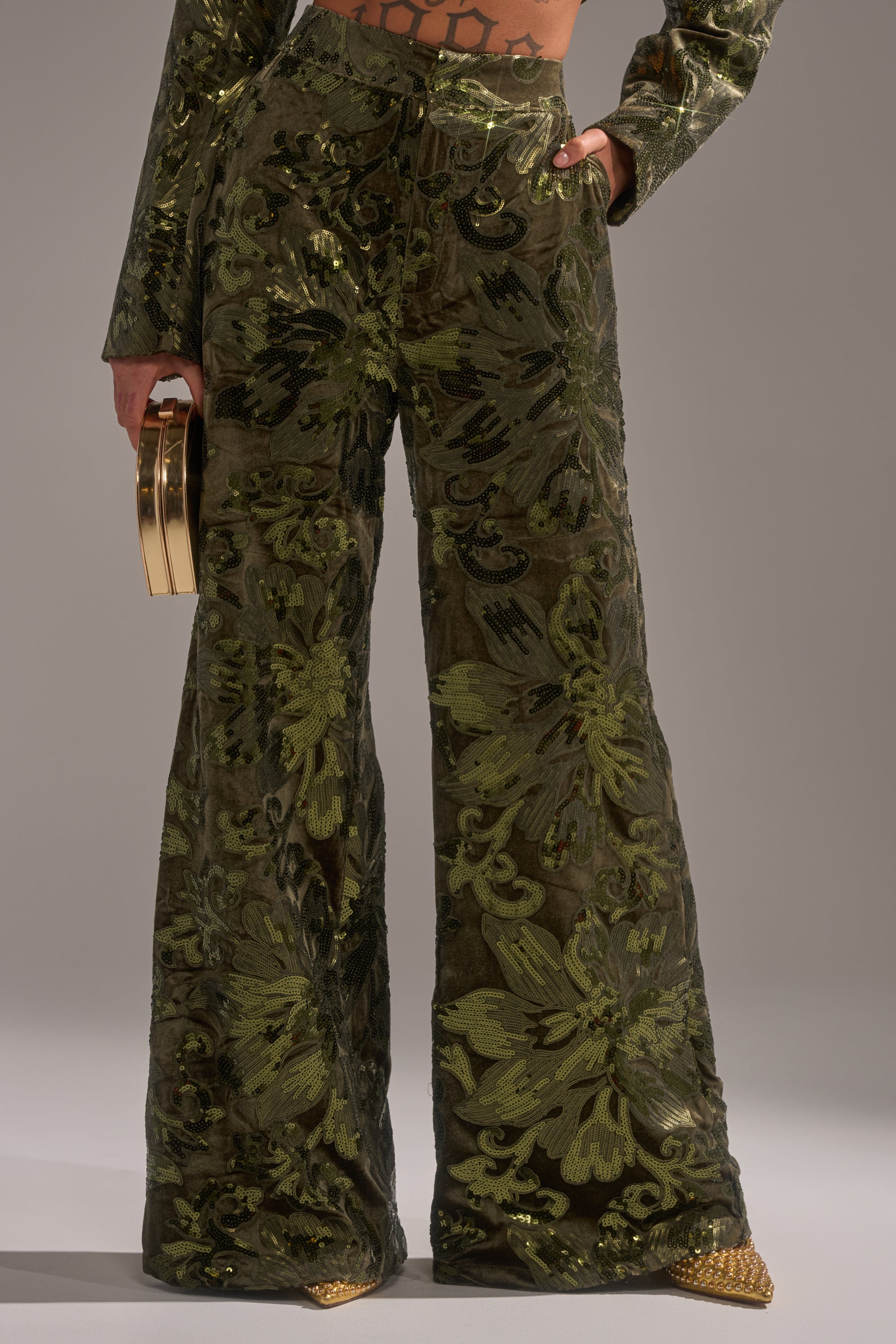 A person wears the AURORA SEQUIN VELVET TROUSER, high-waisted with a green floral, wide-leg silhouette and a shiny textured pattern. They hold a gold clutch and wear pointed gold shoes; only their lower half is visible.