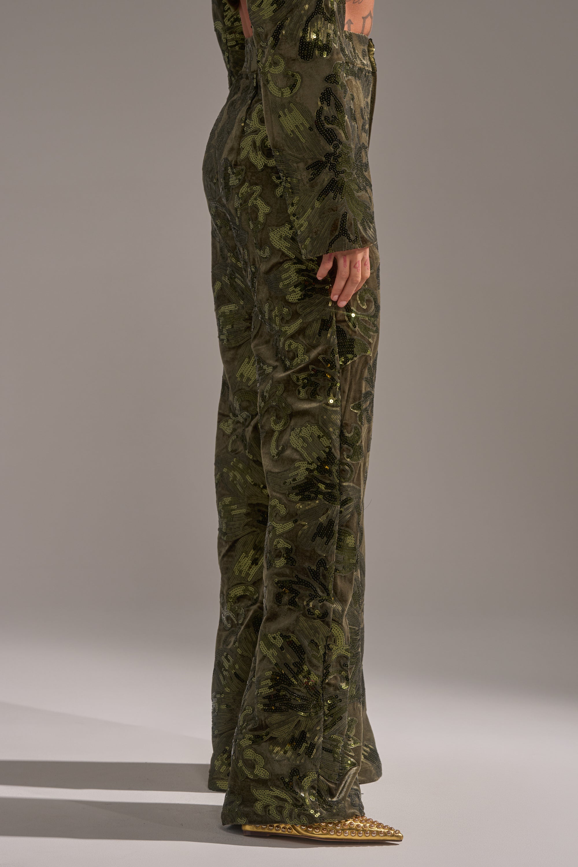 A person standing sideways wears the AURORA SEQUIN VELVET TROUSER, featuring a green and brown camouflage pattern, paired with gold pointed shoes against a plain gray background.