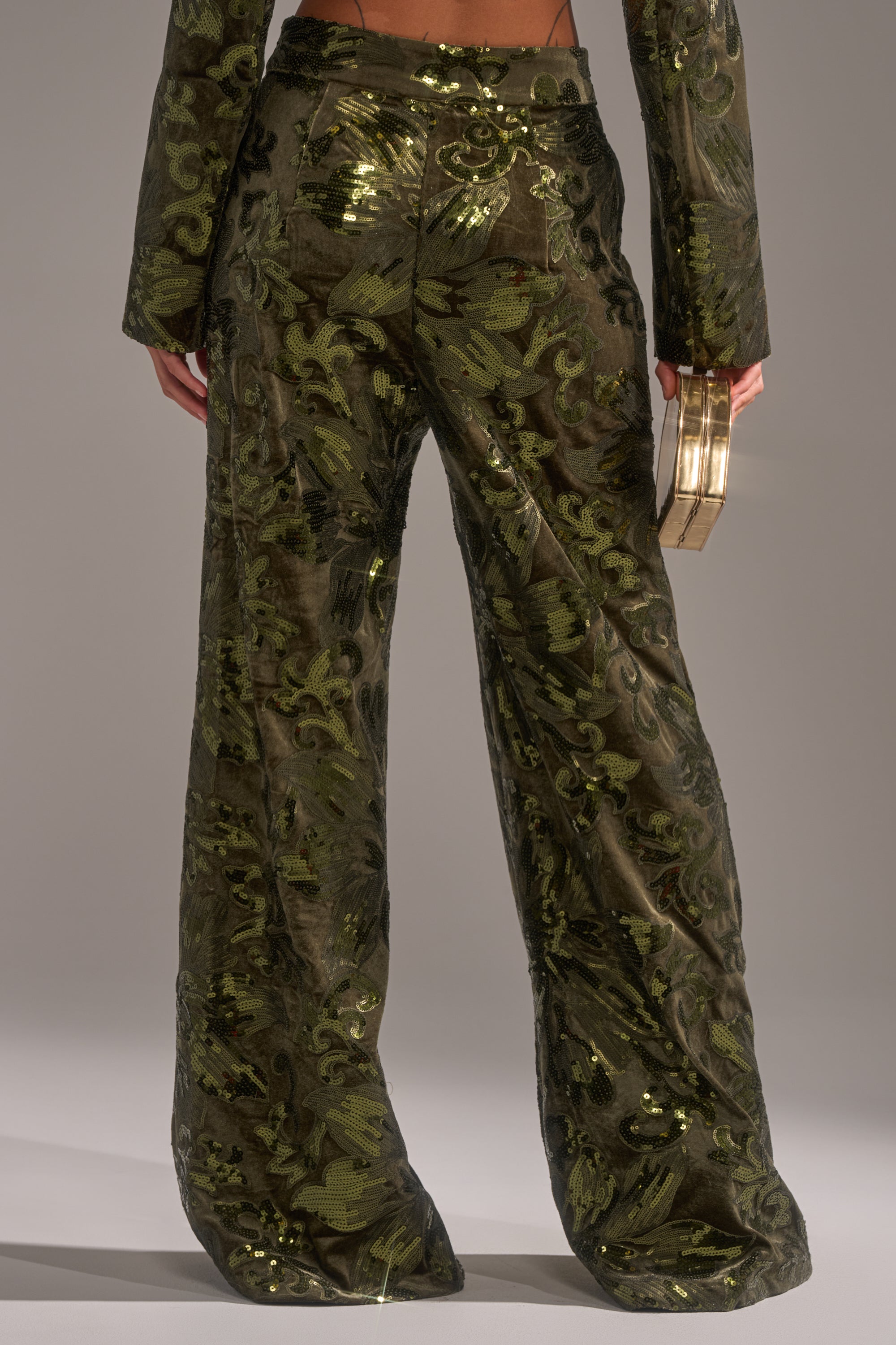 Someone is standing with their back to the camera, wearing the AURORA SEQUIN VELVET TROUSER and a matching jacket in dark green floral-patterned sequins, holding a gold geometric clutch in their left hand.