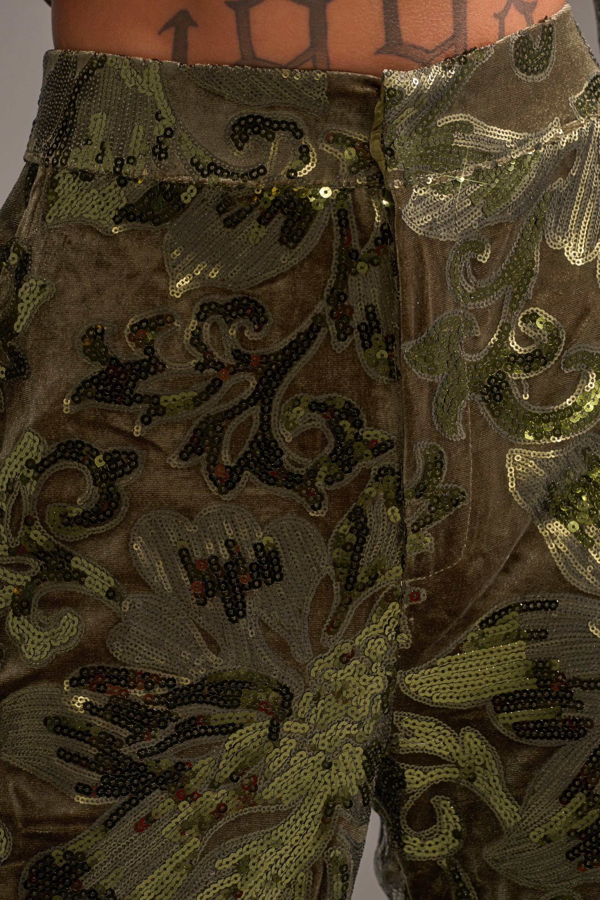 Close-up of the AURORA SEQUIN VELVET TROUSER in olive green, featuring a shimmering floral sequin pattern on high-waisted velvet, with the waistband and wearer’s tattooed lower torso visible.