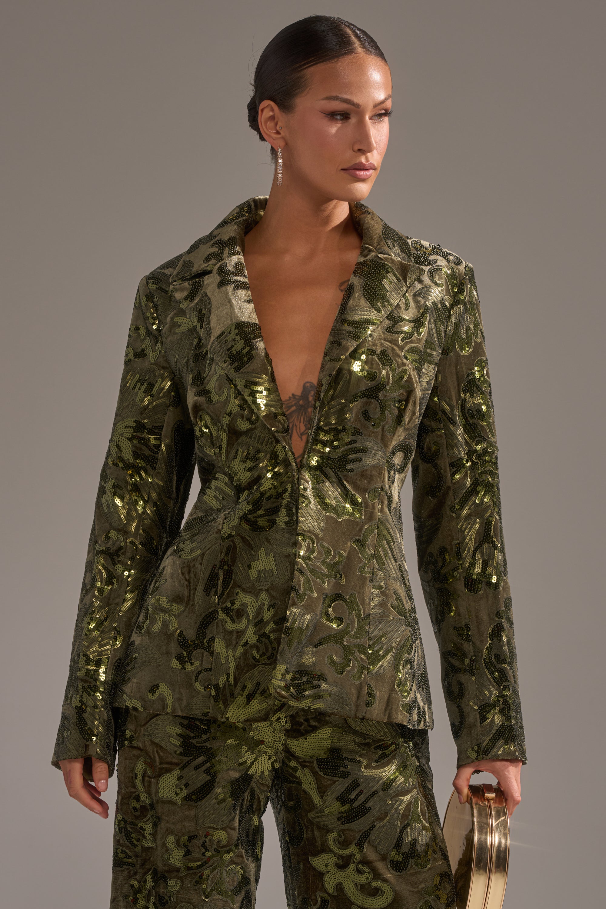 A woman wearing the AURORA SEQUIN VELVET BLAZER and matching floral pants stands against a neutral background, holding a small gold clutch and looking to the side with her hair neatly pulled back.