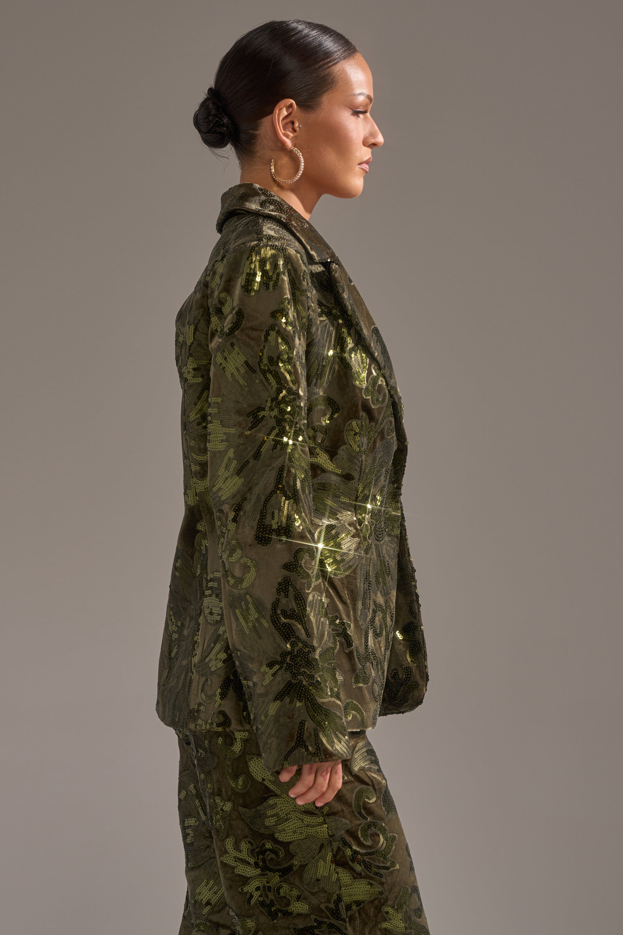 A woman in profile stands against a neutral background, dressed in an olive green, patterned AURORA SEQUIN VELVET BLAZER with matching pants. Her dark hair is styled in a bun, accessorized with large hoop earrings.