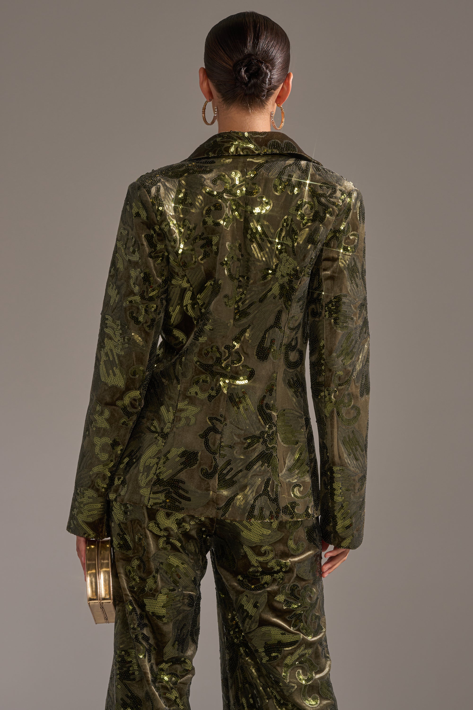 A person with dark hair in a sleek bun, facing away from the camera against a plain gray background, wears the AURORA SEQUIN VELVET BLAZER and holds a small metallic box-shaped purse.