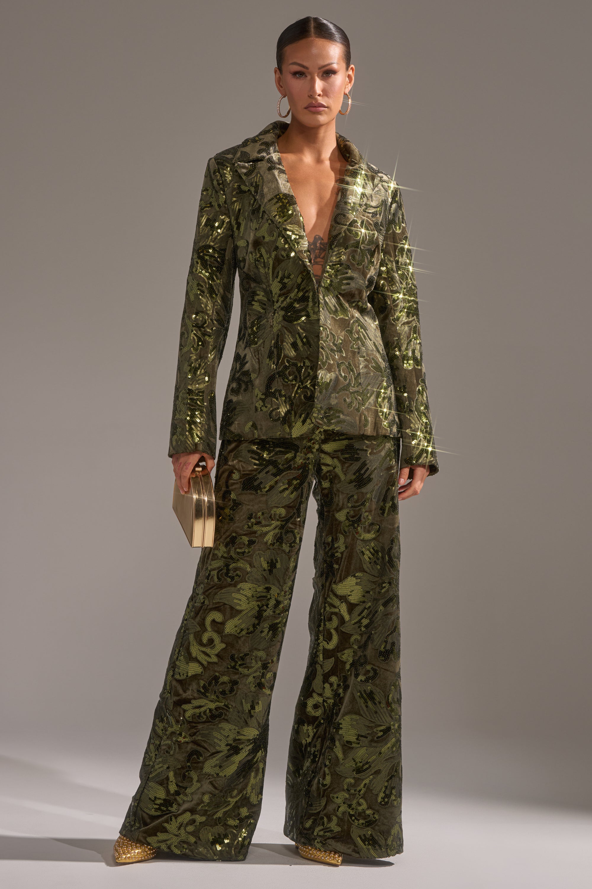 A person stands confidently in a shiny patterned green pantsuit with wide-leg trousers and the AURORA SEQUIN VELVET BLAZER, accessorized with a gold clutch, gold pointed shoes, hoop earrings, and slicked-back hair.