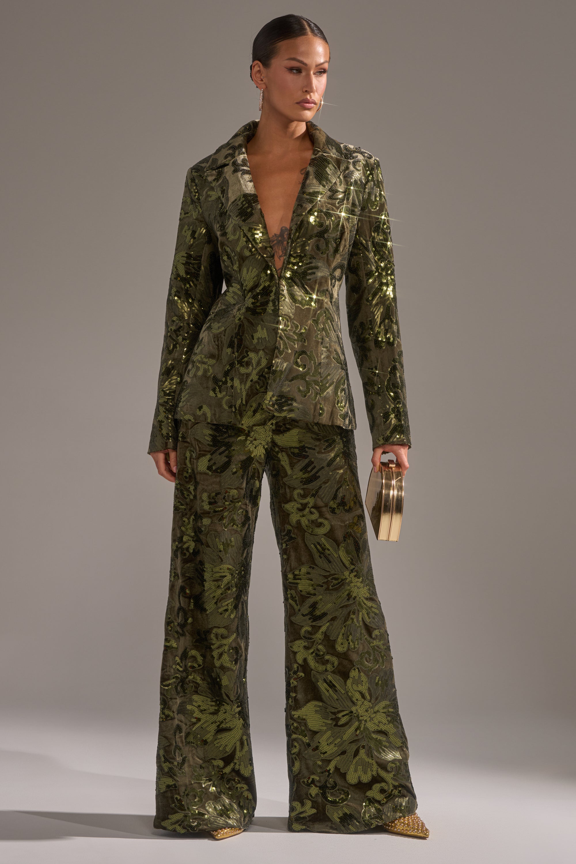 A woman stands confidently in the AURORA SEQUIN VELVET BLAZER with a deep V-neckline, paired with a small gold clutch and gold pointed-toe shoes, set against a neutral background.