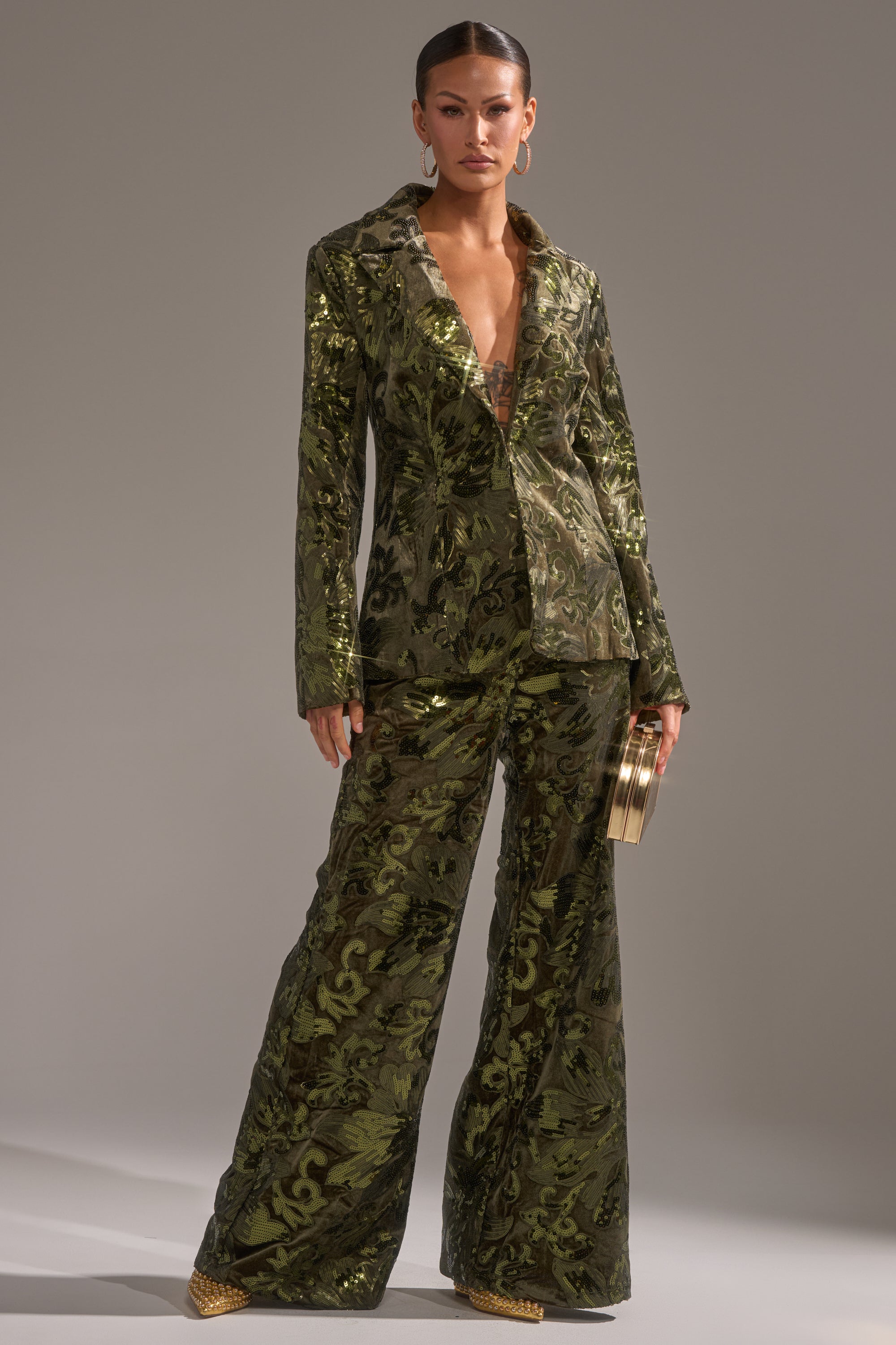 A woman stands confidently in the AURORA SEQUIN VELVET BLAZER and matching wide-leg pants with a baroque motif, accessorized with statement earrings and a gold clutch, posing against a neutral backdrop.